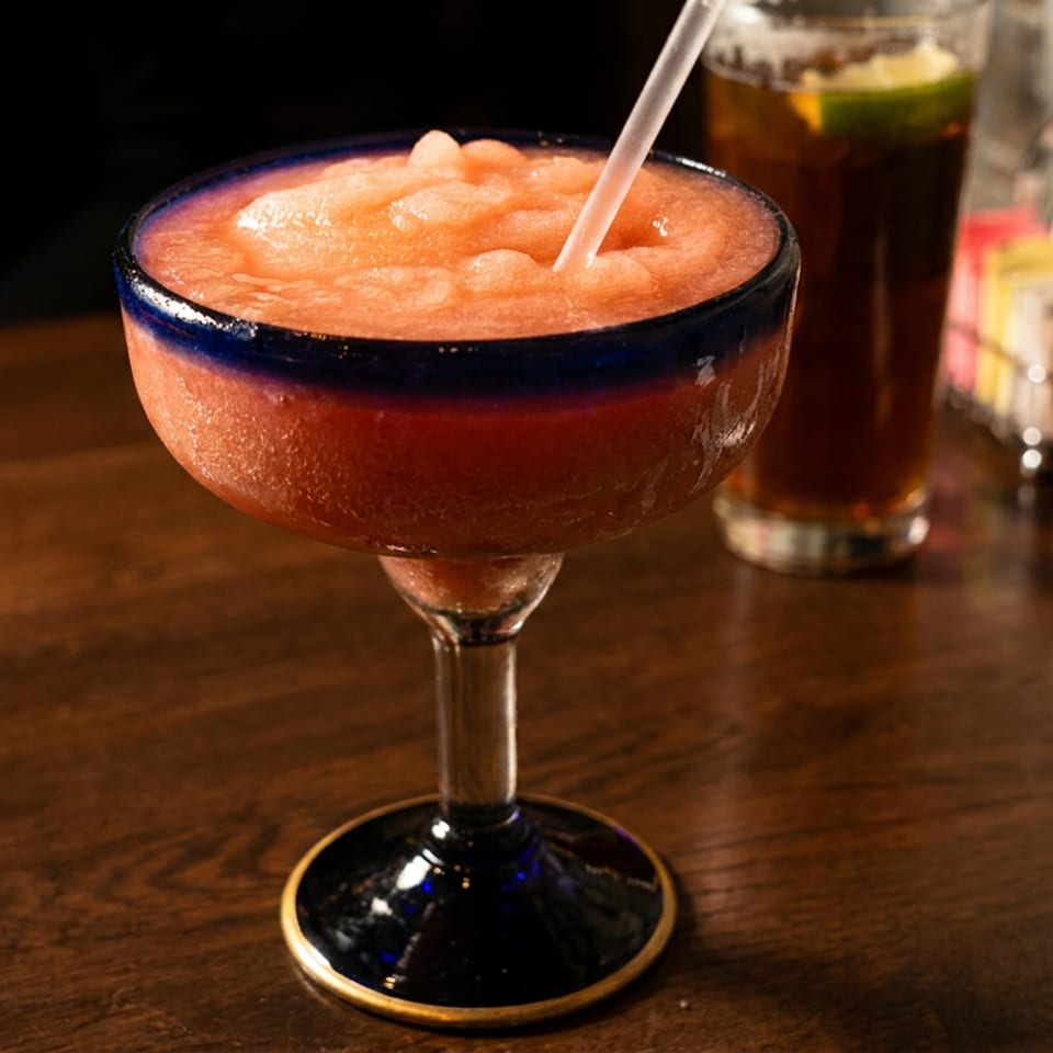 A vibrant red frozen margarita in a blue-rimmed glass with a straw on a wooden table.